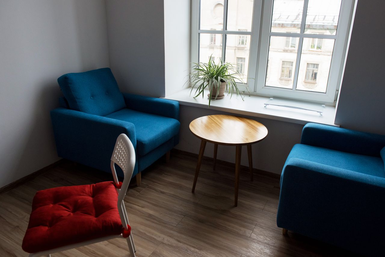 Comfortable blue armchairs with soft cushions and round wooden table near window with flowerpot in room with white walls and wooden floor